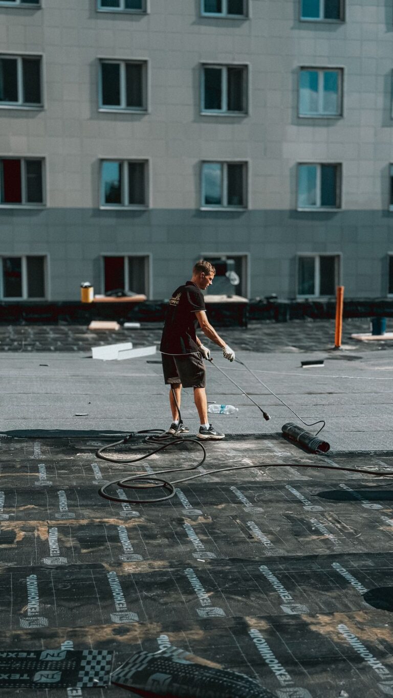 A construction worker uses a torch to apply roofing material on a city building under sunlight.